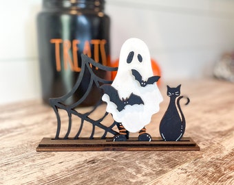 Pode incluir: Uma decoração de Halloween de madeira com um fantasma branco com morcegos pretos, um gato preto e uma teia de aranha preta. A decoração está sobre uma base de madeira castanho.