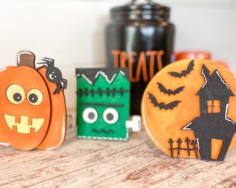 May include: Three Halloween-themed wooden decorations. A pumpkin with a spider on top, a green Frankenstein monster, and a black silhouette of a house with bats flying around a full moon.