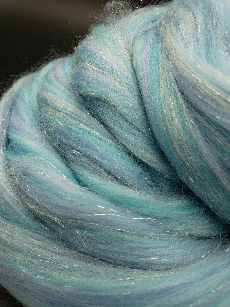 K&ouml;nnte beinhalten: Nahaufnahme eines B&uuml;ndels blauer und wei&szlig;er Wollroving mit silbernen Schimmerf&auml;den. Der Roving ist weich und flauschig, und die Farben sind leuchtend.