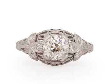 May include: A silver ring with a large round diamond in the center and smaller diamonds on the band. The band is decorated with a delicate filigree design.