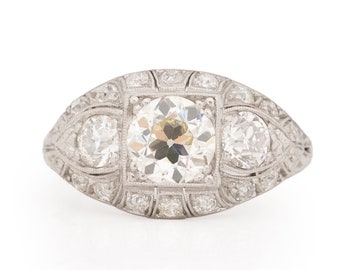 Könnte beinhalten: Ein Vintage-Verlobungsring aus Platin mit einem großen runden Diamanten in der Mitte, der von kleineren Diamanten umgeben ist. Der Ring hat ein zartes, filigranes Design.