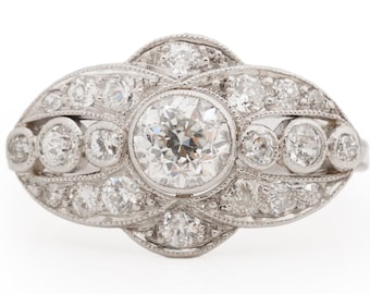 Könnte beinhalten: Ein Vintage-Diamant-Verlobungsring mit einem großen, runden Mittelstück, das von kleineren Diamanten umgeben ist. Der Ring ist in Weißgold gefasst und hat ein zartes, filigranes Design.