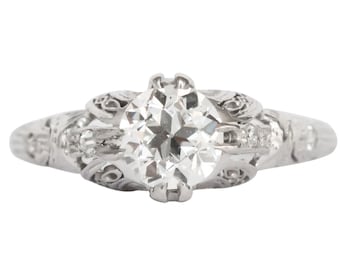 Könnte beinhalten: Ein Verlobungsring im Vintage-Stil mit einem großen, rund geschliffenen Diamanten als Mittelpunkt. Der Ring weist aufwendige Filigranarbeiten entlang des Bandes und um den zentralen Stein auf, mit kleineren Akzentdiamanten. Das Metall scheint Weißgold oder Platin zu sein.