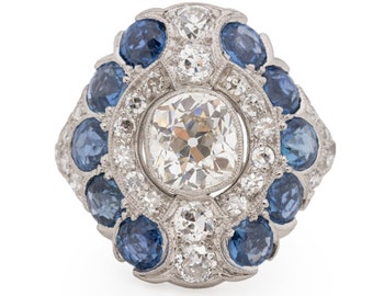 May include: A platinum ring featuring a large, cushion-cut diamond surrounded by a halo of smaller diamonds and blue sapphires.