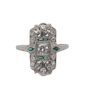 May include: A silver ring with a rectangular face, featuring a central square diamond, surrounded by smaller diamonds and emerald accents. The ring has an Art Deco design with intricate detailing.