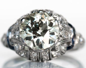 Puede incluir: Un anillo de compromiso de platino vintage con un gran diamante redondo de talla brillante rodeado de diamantes más pequeños y zafiros azules.