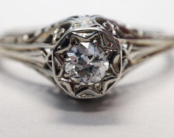 May include: A silver engagement ring with a delicate filigree design and a round, clear diamond in the center.