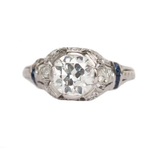 May include: A vintage engagement ring with a large, round, clear diamond center stone surrounded by smaller diamonds and blue sapphires. The ring is set in platinum and has a delicate, intricate design.