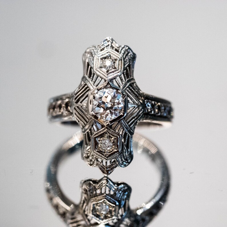 May include: A silver ring with a large, clear diamond in a geometric setting. The ring is surrounded by smaller diamonds and has a delicate, intricate design.