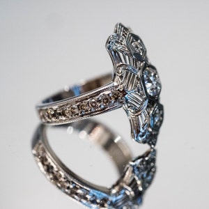 May include: A silver ring with a large, clear diamond in the center and smaller diamonds surrounding it. The band of the ring is also set with smaller diamonds.