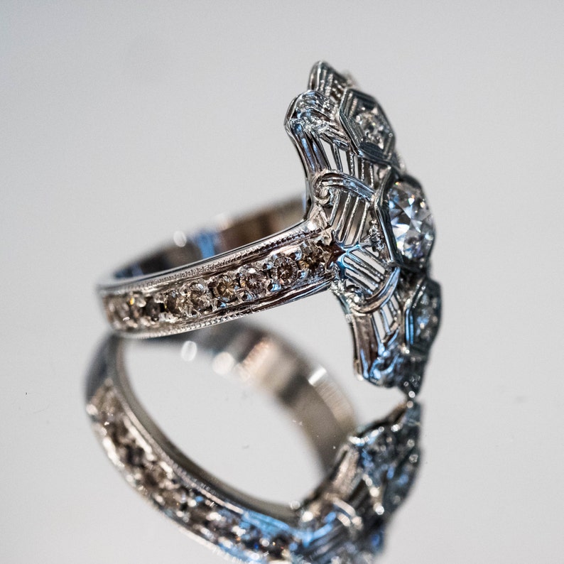 May include: A silver ring with a large, clear diamond in the center and smaller diamonds surrounding it. The band of the ring is also set with smaller diamonds.