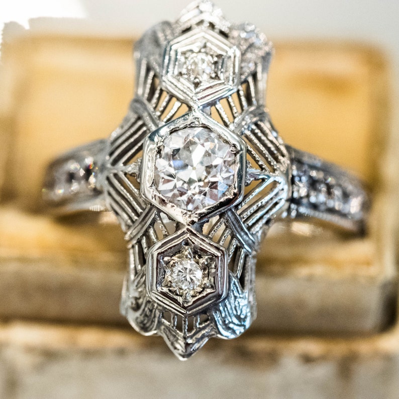 May include: A silver ring with a large, round, clear diamond in the center. The ring has a geometric design with smaller diamonds on the sides.