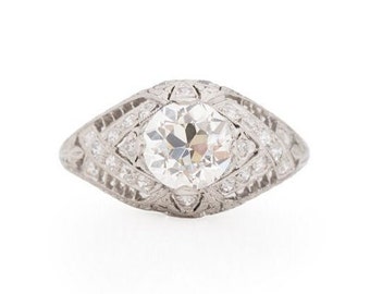 May include: A vintage platinum engagement ring with a round brilliant cut diamond surrounded by smaller diamonds. The ring has a delicate, intricate design.