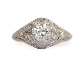 May include: A vintage-style engagement ring with a large, round, clear diamond center stone. The ring is set in a white metal with intricate filigree and small accent diamonds. The ring is set against a white background.