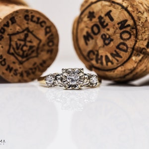 May include: A gold ring with a large, round, clear diamond in the center and smaller diamonds on either side. The ring is sitting on a white surface in front of two champagne bottle corks.