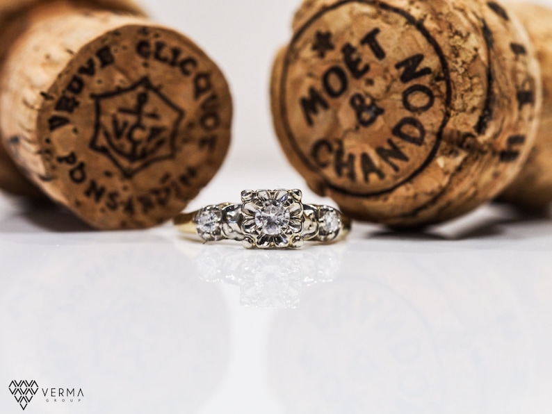 May include: A gold ring with a large, round, clear diamond in the center and smaller diamonds on either side. The ring is sitting on a white surface in front of two champagne bottle corks.