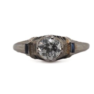 May include: A silver ring with a round, clear diamond center stone and two small blue sapphire accent stones.