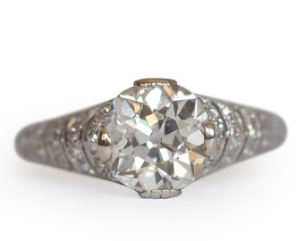 Puede incluir: Un anillo de compromiso vintage con un gran diamante redondo de talla brillante engastado en una banda de platino. La banda está decorada con un delicado diseño grabado.
