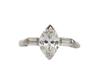 Puede incluir: Un anillo de plata con un diamante de forma marquesa en el centro y dos diamantes más pequeños de forma baguette a cada lado.