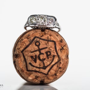 May include: A silver engagement ring with a large center diamond and two smaller diamonds on either side, sitting on top of a champagne cork with the letters "VCP" and an anchor design.