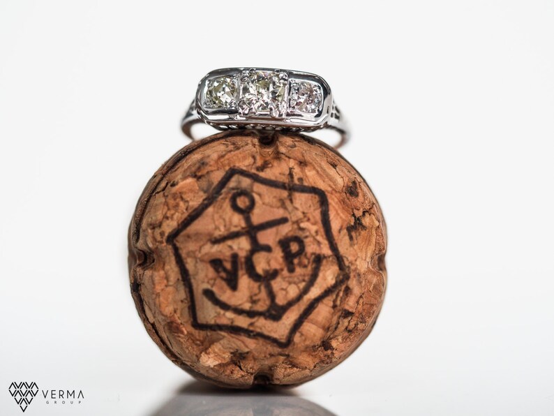 May include: A silver engagement ring with a large center diamond and two smaller diamonds on either side, sitting on top of a champagne cork with the letters "VCP" and an anchor design.