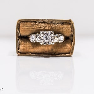 May include: A silver ring with a large, round, clear diamond in the center and smaller diamonds on either side. The ring is in a brown velvet box.