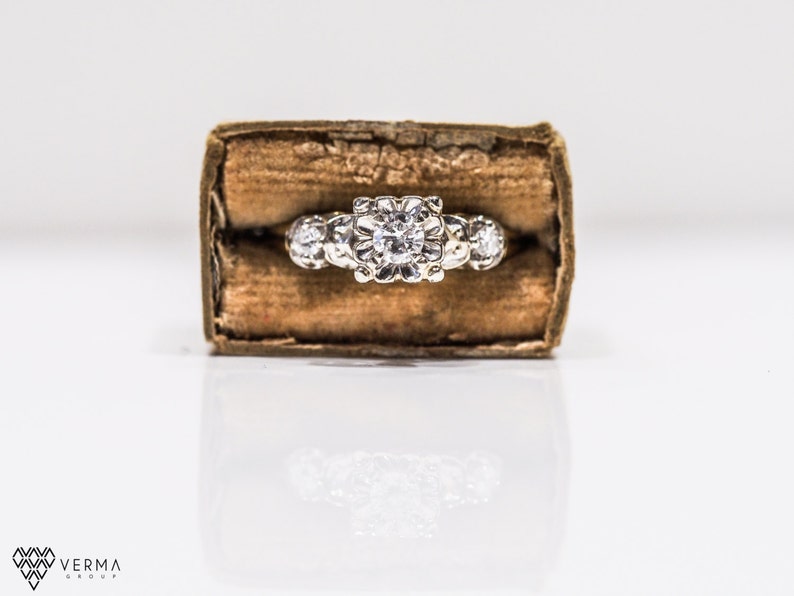 May include: A silver ring with a large, round, clear diamond in the center and smaller diamonds on either side. The ring is in a brown velvet box.