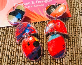 May include: A pair of colorful, abstract earrings with a red, blue, and black design. The earrings are made of resin and have a clear, glossy finish. The earrings are attached to a silver hook.