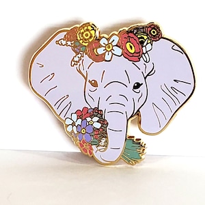 May include: A purple enamel pin of an elephant wearing a flower crown and holding a bouquet of flowers. The elephant has a gold outline and the flowers are in shades of pink, red, yellow, blue, and green.