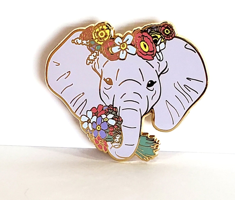May include: A purple enamel pin of an elephant wearing a flower crown and holding a bouquet of flowers. The elephant has a gold outline and the flowers are in shades of pink, red, yellow, blue, and green.