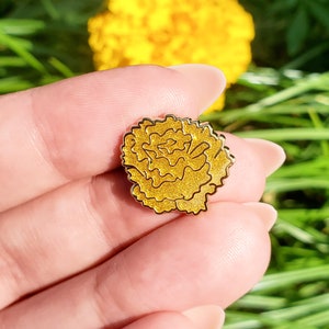 May include: A gold-colored enamel pin shaped like a marigold flower. The pin has a textured surface that resembles petals.