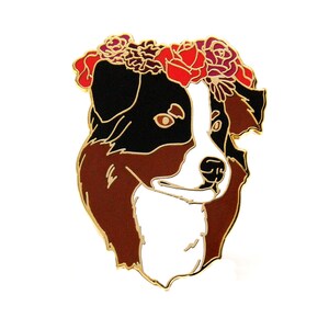 May include: A colorful enamel pin featuring a dog's head with a floral crown. The dog has a black and white face with brown and gold accents. The flower crown is red, burgundy, and pink.