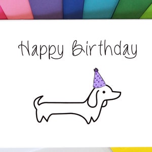May include: A white card with the text "Happy Birthday" written in black cursive. A black and white illustration of a dachshund wearing a purple party hat is below the text.