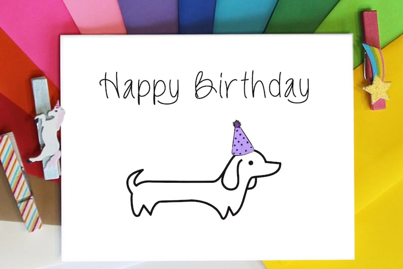 May include: A white card with the text "Happy Birthday" written in black cursive. A black and white illustration of a dachshund wearing a purple party hat is below the text.