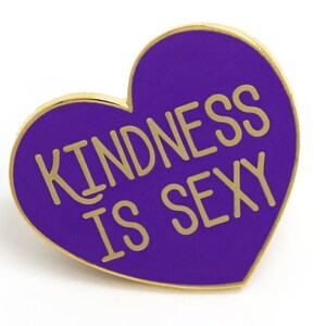 K&ouml;nnte beinhalten: Herzf&ouml;rmiger Emaille-Ansteckpin mit goldenem Rand und dem Schriftzug "KINDNESS IS SEXY" in goldfarbenen Buchstaben auf lila Hintergrund. Der Pin ist ein dekoratives Accessoire.