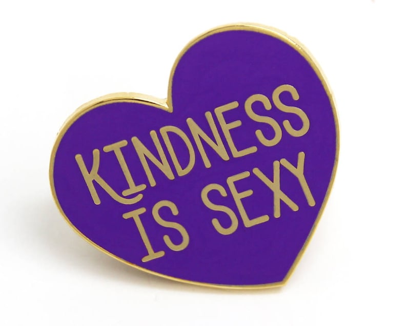 K&ouml;nnte beinhalten: Herzf&ouml;rmiger Emaille-Ansteckpin mit goldenem Rand und dem Schriftzug "KINDNESS IS SEXY" in goldfarbenen Buchstaben auf lila Hintergrund. Der Pin ist ein dekoratives Accessoire.