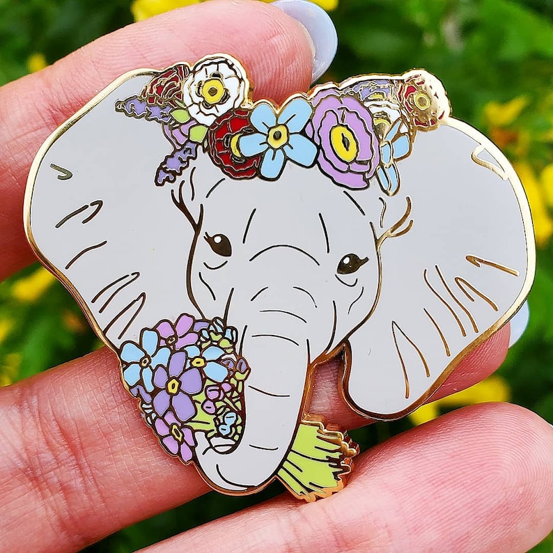 May include: A gold-tone enamel pin featuring a gray elephant with a floral crown and a bouquet of flowers in its trunk. The elephant has a light gray body and a white trunk. The flowers are pink, purple, blue, and yellow.