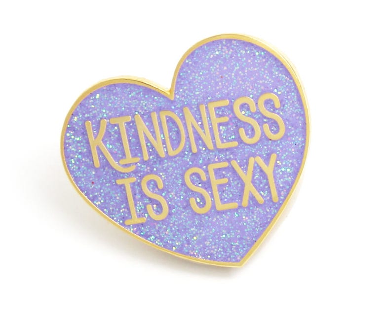 K&ouml;nnte beinhalten: Herzf&ouml;rmige Emaille-Anstecknadel mit glitzerndem, lavendelfarbenem Hintergrund und goldfarbenem Rand. Die Anstecknadel tr&auml;gt den Text "KINDNESS IS SEXY" in goldfarbenen Buchstaben. Die Anstecknadel ist ein dekoratives Accessoire.