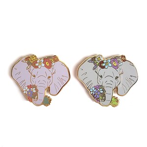 May include: Two enamel pins featuring an elephant with a floral crown and a bouquet of flowers. One elephant is light purple and the other is light gray.