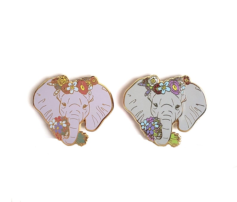 May include: Two enamel pins featuring an elephant with a floral crown and a bouquet of flowers. One elephant is light purple and the other is light gray.