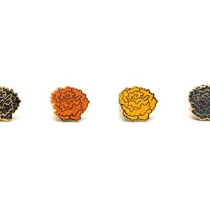 May include: Four flower-shaped enamel pins with gold-coloured borders and glittery centres. The pins are in dark blue, orange, yellow, and dark blue. They are arranged on a white background.