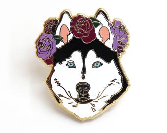 May include: A gold-tone enamel pin featuring a white husky dog with black fur around its eyes and ears. The dog is wearing a purple and burgundy flower crown.
