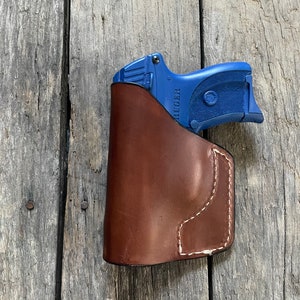 Może przedstawiać: Brązowa sk&oacute;rzana kabura z niebieskim pistoletem w środku. Pistolet ma napis "RUGER" nadrukowany na boku.