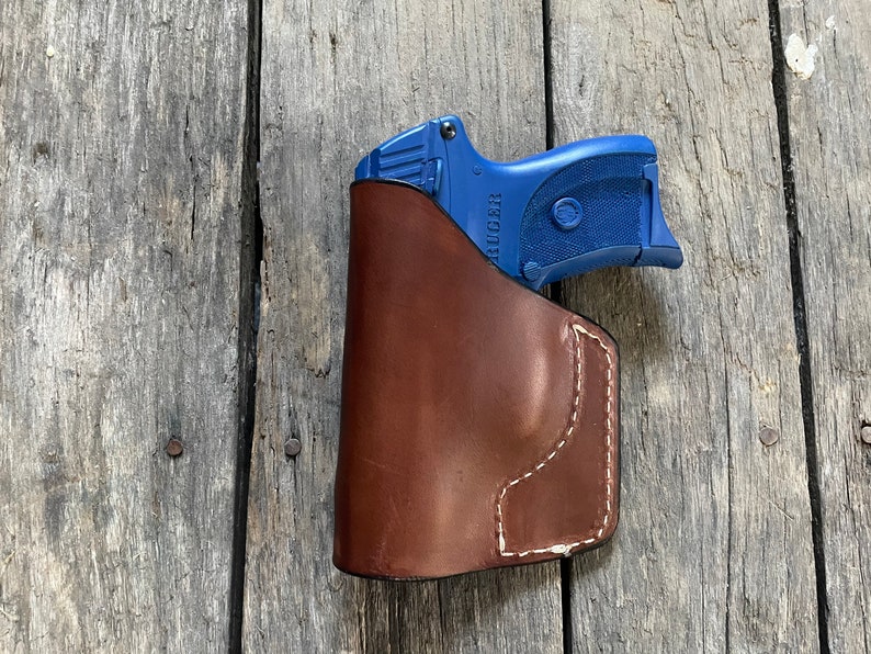 Może przedstawiać: Brązowa sk&oacute;rzana kabura z niebieskim pistoletem w środku. Pistolet ma napis "RUGER" nadrukowany na boku.