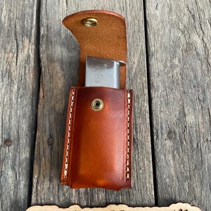 Może przedstawiać: Brązowy skórzany futerał na magazynki z klapką i zatrzaskiem. Futerał jest otwarty, a wewnątrz znajduje się srebrny magazynek. Futerał leży na drewnianej powierzchni. @HarbourMercantile