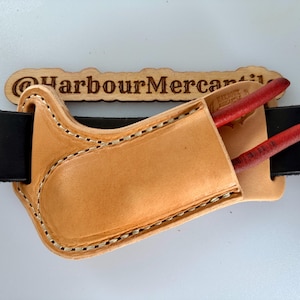 May include: A tan leather tool holster with visible stitching, threaded onto a black leather belt. The holster contains a pair of pliers with red handles. A wooden sign with the laser-engraved text @HarbourMercantile is positioned behind the belt.