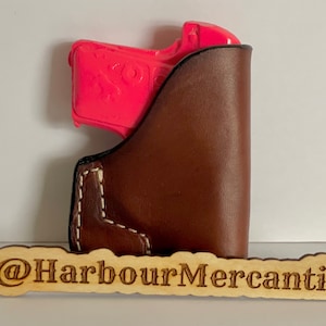 May include: A brown leather holster with a pink plastic toy gun inside. The holster has white stitching and a curved shape. The image includes the text "@HarbourMercantile".