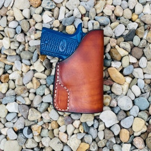 May include: A blue handgun in a brown leather holster. The holster has a stitched design and is sitting on a bed of small, gray rocks.