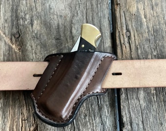 Peut inclure: Un étui à couteau en cuir marron foncé avec des coutures blanches apparentes, conçu pour se glisser sur une ceinture en cuir beige clair. Un couteau pliant avec une mitre en laiton et un manche noir est partiellement inséré dans l'étui.