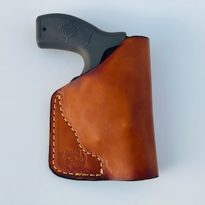 Może przedstawiać: Brązowa skórzana kabura z pikowanym wzorem i napisem "Barbour Leather" na dole. Czarny pistolet jest częściowo widoczny w kaburze.